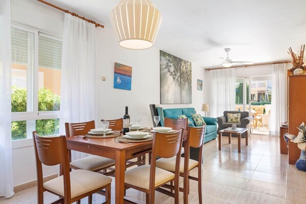 A dining area with a wooden table set for four, a teal sofa, and a ceiling fan. - Villa El Olivo - Three Bedroom Villa, Sleeps 6 (Pollença)