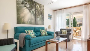 Ein Wohnzimmer mit einem türkisfarbenen Sofa, einem grauen Sessel und einem Couchtisch. An der Wand hängt ein großes Gemälde von Bäumen, eine Lampe ist vorhanden und es gibt einen Blick auf den Außenbalkon.