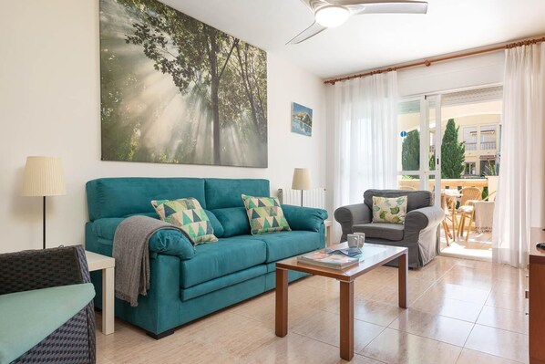 Una sala de estar con un sofá color turquesa, un sillón gris y una mesa de centro. Se observa un cuadro grande con árboles en la pared, una lámpara y vistas al balcón exterior.