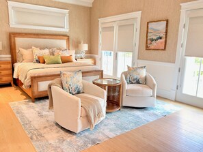 Une chambre à coucher avec un grand lit, deux fauteuils, une petite table ronde et une vue sur l’extérieur par une fenêtre.