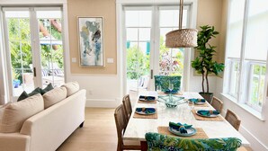 A dining area with a table set for four, surrounded by wicker chairs, a white sofa, and a large window with a view of greenery. - Luxury Windsor, Key West Retreat- Old Town ; Pool, Gated Parking, Walk to Duval (Key West)