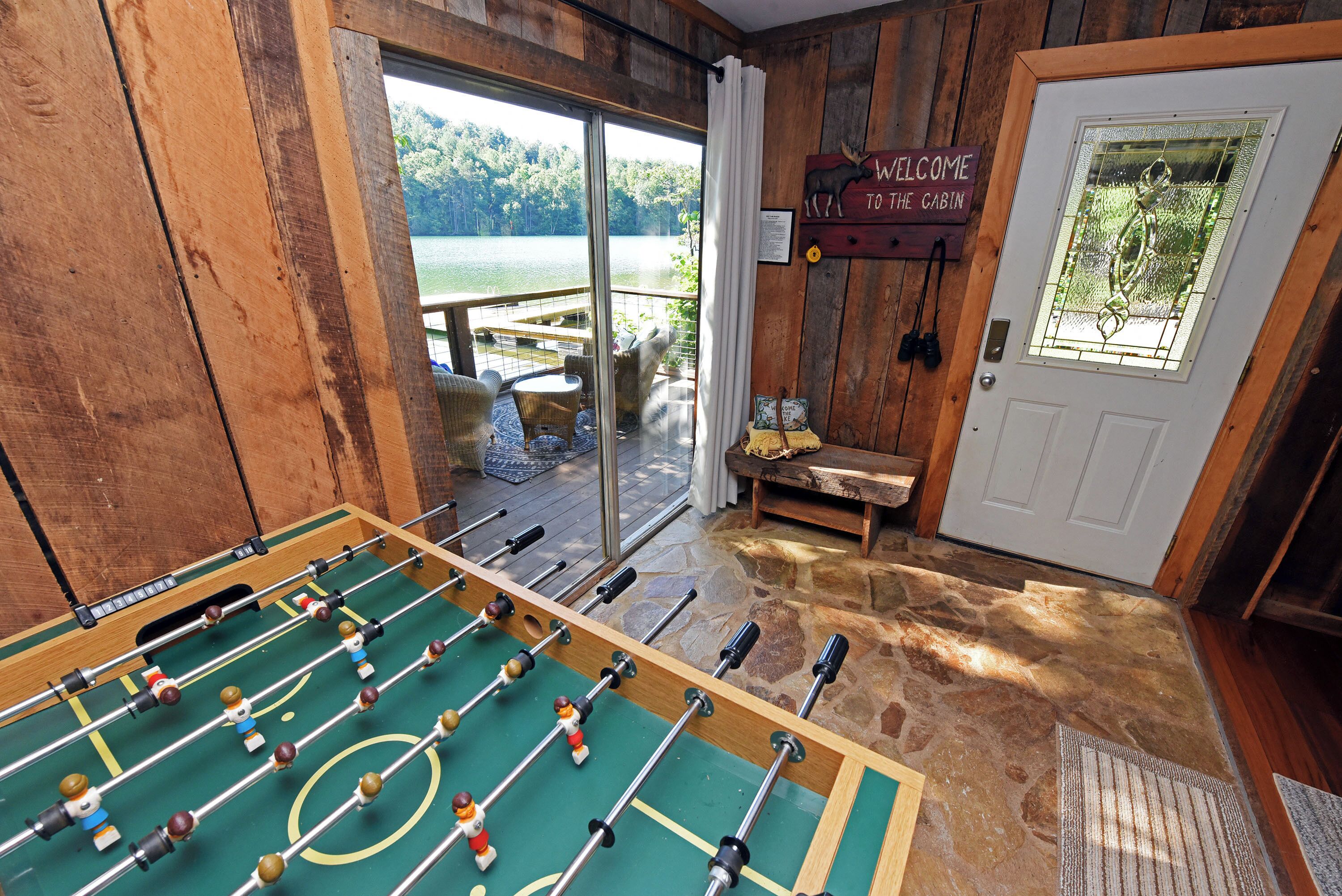 A room with a foosball table, a wooden door with a welcome sign, and a view of a lake through large glass doors.