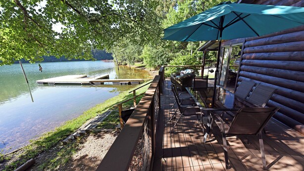 Une terrasse en bois offrant une vue sur un lac, avec un quai et un espace salon couvert agrémenté d’un parasol.