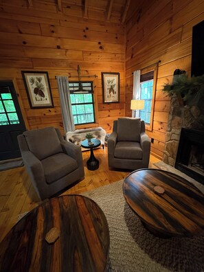 A cozy cabin interior with wooden walls, two armchairs, a round coffee table, a fireplace, and framed botanical prints on the walls. - Tranquil 1-bedroom 4 Person Cabin in charming Blue Ridge Cabin Community (Blue Ridge)