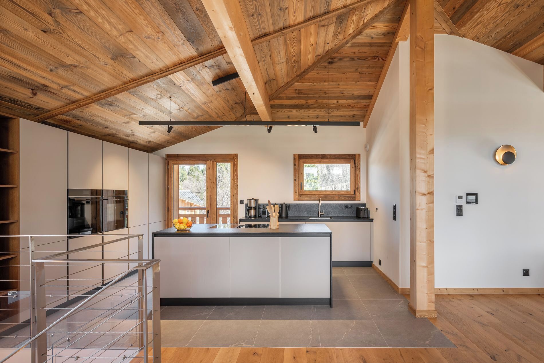 Een moderne keuken met een centraal eiland, houten balken in het plafond en uitzicht op de omgeving door een raam.