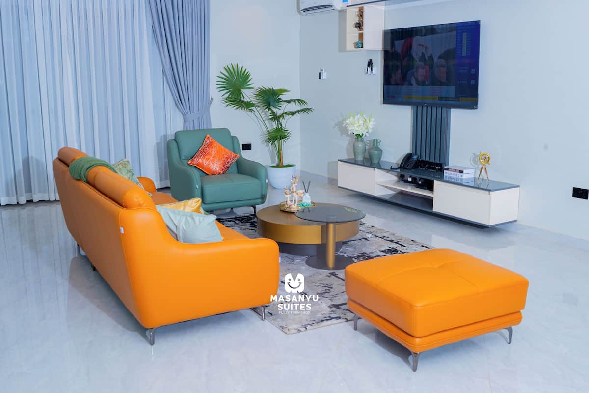 A modern living room with a sofa, armchair, ottoman, and a coffee table. There is a TV mounted on the wall and a potted plant.