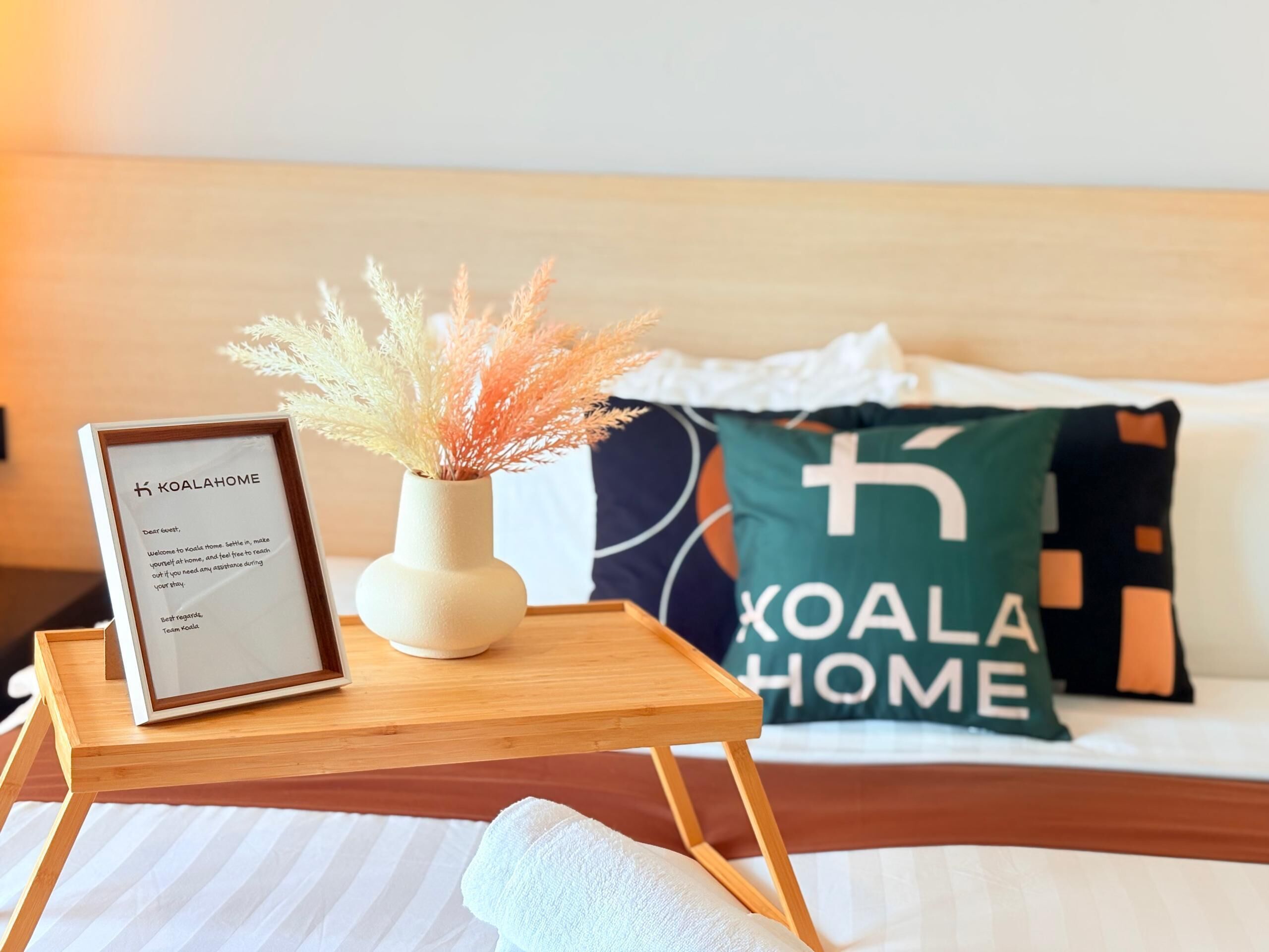 A bed with a pillow featuring "KOALA HOME" text, a framed welcome message, and a small vase with dried plants on a bedside table.