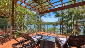 A wooden deck with wicker chairs and a glass table, overlooking a lake with palm trees and a distant mountain. - Dorathee Cottage - Pet Friendly - Absolute Waterfront (Gunderman)