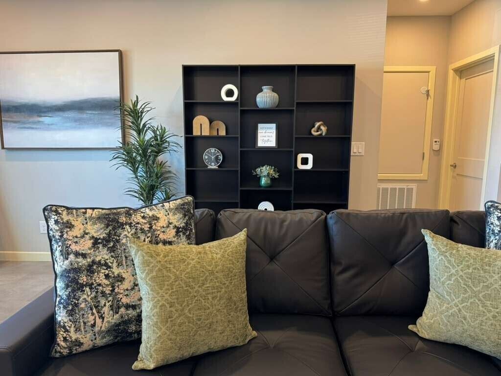 Una sala de estar con un sofá de cuero oscuro, dos cojines estampados, un mueble negro con varios objetos decorativos, un cuadro en la pared y una planta en maceta.