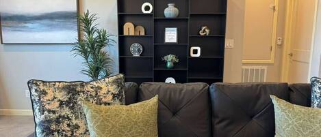 Una sala de estar con un sofá de cuero oscuro, dos cojines estampados, un mueble negro con varios objetos decorativos, un cuadro en la pared y una planta en maceta.