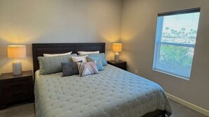 Une chambre à coucher avec un grand lit, deux tables de chevet avec des lampes, un tapis à motifs et une fenêtre avec des stores.