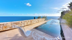 Piscina infinita com muro de pedra e vista para o oceano. Duas cadeiras de metal branco e uma pequena mesa.