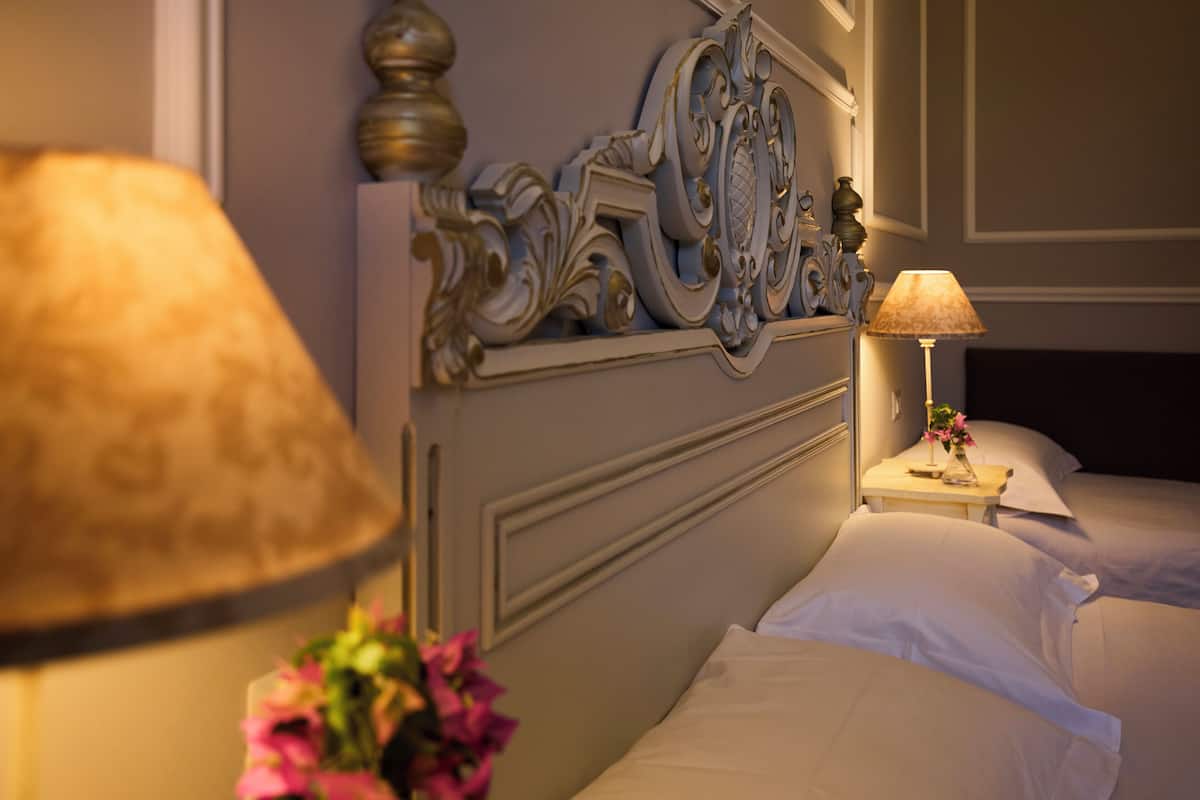 A hotel room with a classic design, featuring a decorative headboard, two beds, a bedside table with a lamp, and a vase with flowers.