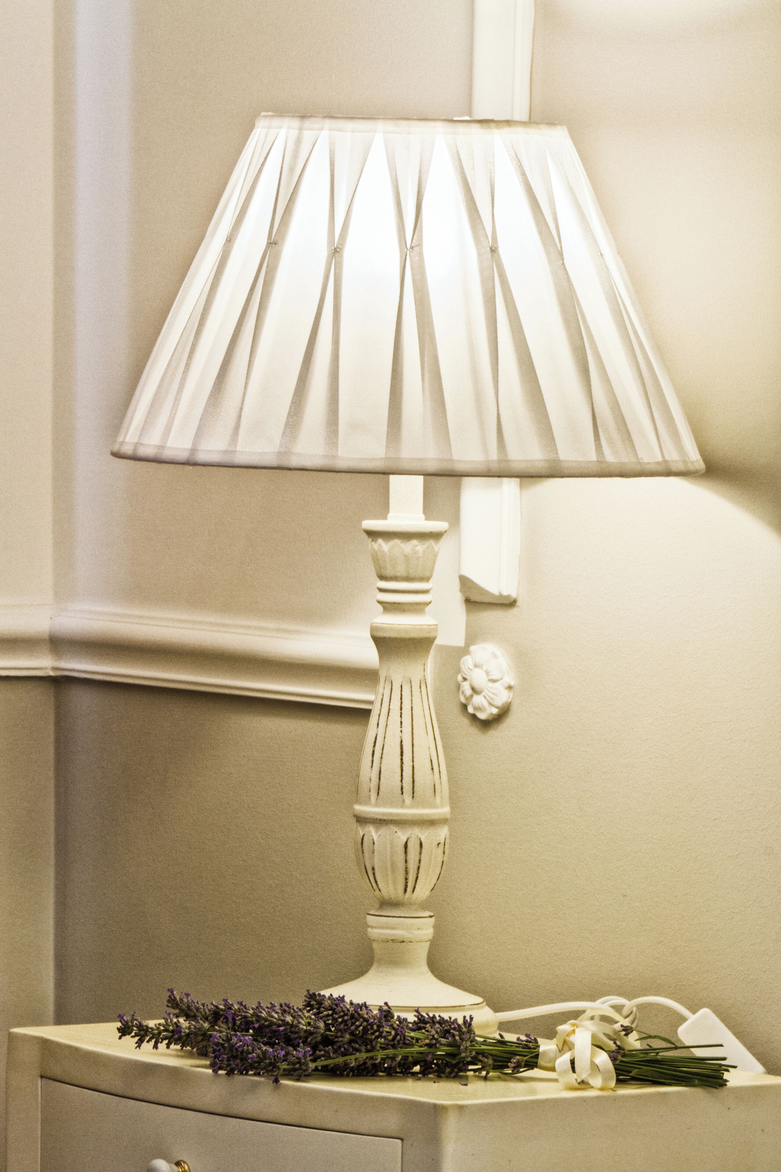 Une table de chevet avec une lampe blanche, un petit objet décoratif et un bouquet de fleurs de lavande.
