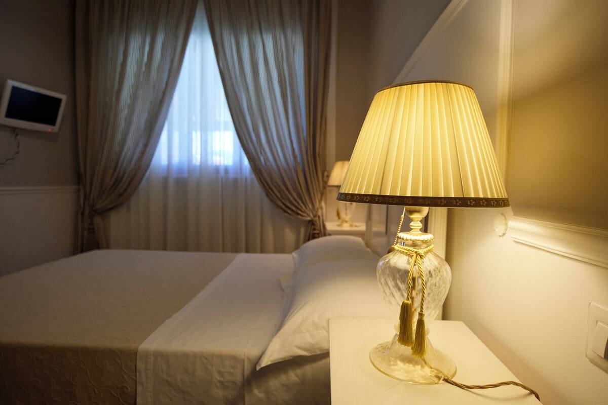 A hotel room with a bed, a bedside lamp, a TV on the wall, and a window with curtains.