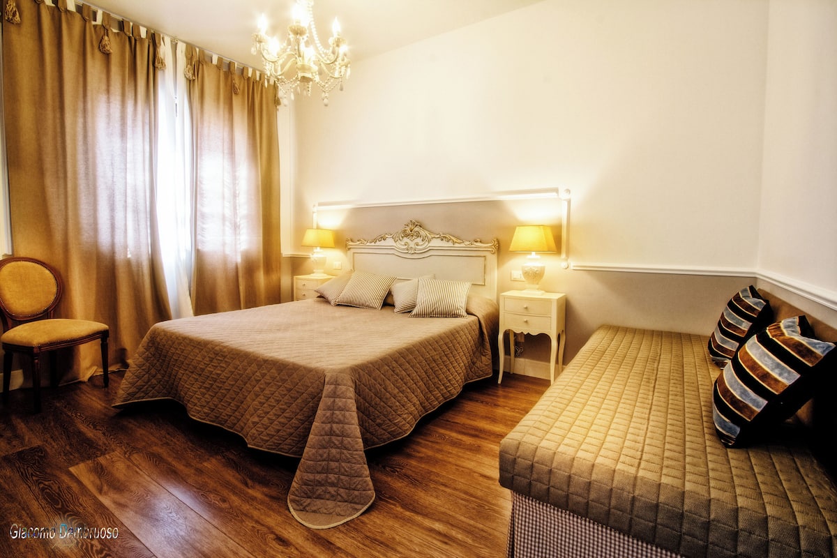 A hotel room with a large bed, two bedside tables with lamps, a chair, and a bench with striped cushions.