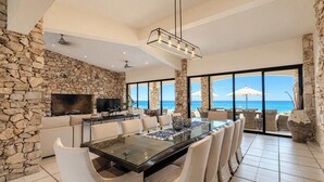 A spacious dining area with a long table and multiple chairs, a stone accent wall, a flat-screen TV, and a view of the ocean. - Oceanfront Villa · Heated Pool, Fire Pit & Beach (San José del Cabo)
