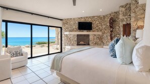A bedroom with a large bed, a television mounted on the wall, and a stone fireplace. - Oceanfront Villa · Heated Pool, Fire Pit & Beach (San José del Cabo)