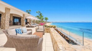 A coastal outdoor seating area with wicker furniture, a stone wall, and a view of the beach and ocean. - Oceanfront Villa · Heated Pool, Fire Pit & Beach (San José del Cabo)