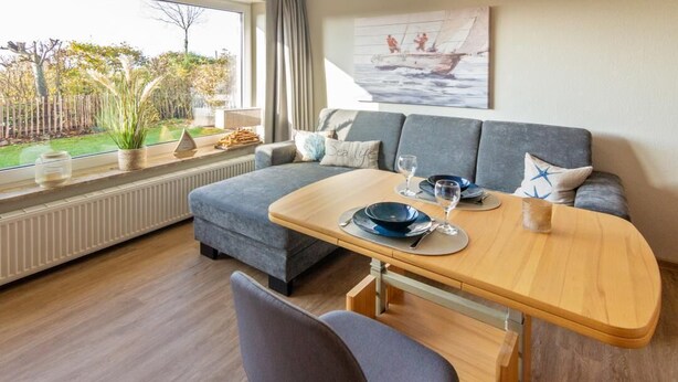 Ein gemütliches Zimmer mit grauem Sofa, einem Holzespeisetisch gedeckt mit Tellern und Gläsern, und Blick auf einen Garten durch ein Fenster.
