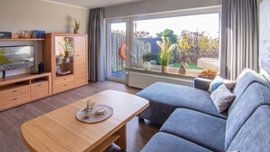 Ein modernes Wohnzimmer mit einer Couch, einem Holztisch und einem Schrank. Ein großes Fenster bietet Blick auf einen Garten.