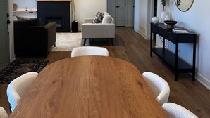 Un salon moderne avec une table à manger en bois, des fauteuils blancs, une cheminée, un téléviseur à écran plat et un lustre.