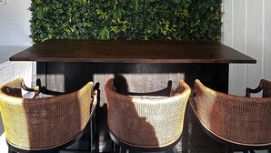 Un coin repas avec une table en bois foncé et des chaises en osier, un mur verdoyant orné d’une enseigne décorative, et un mur blanc en arrière-plan.