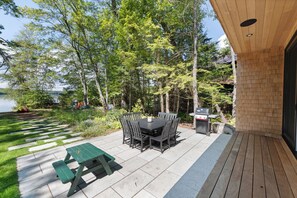 An outdoor patio with a table and chairs, a grill, and a view of a lake surrounded by trees. - NEW! Tilted Pine - Lake Access Modern Home (Center Harbor)