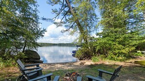 Leirintäalue järven rannalla, jossa on kaksi Adirondack-tyylistä puutarhatuolia, nuotiopaikka ja kanootti.