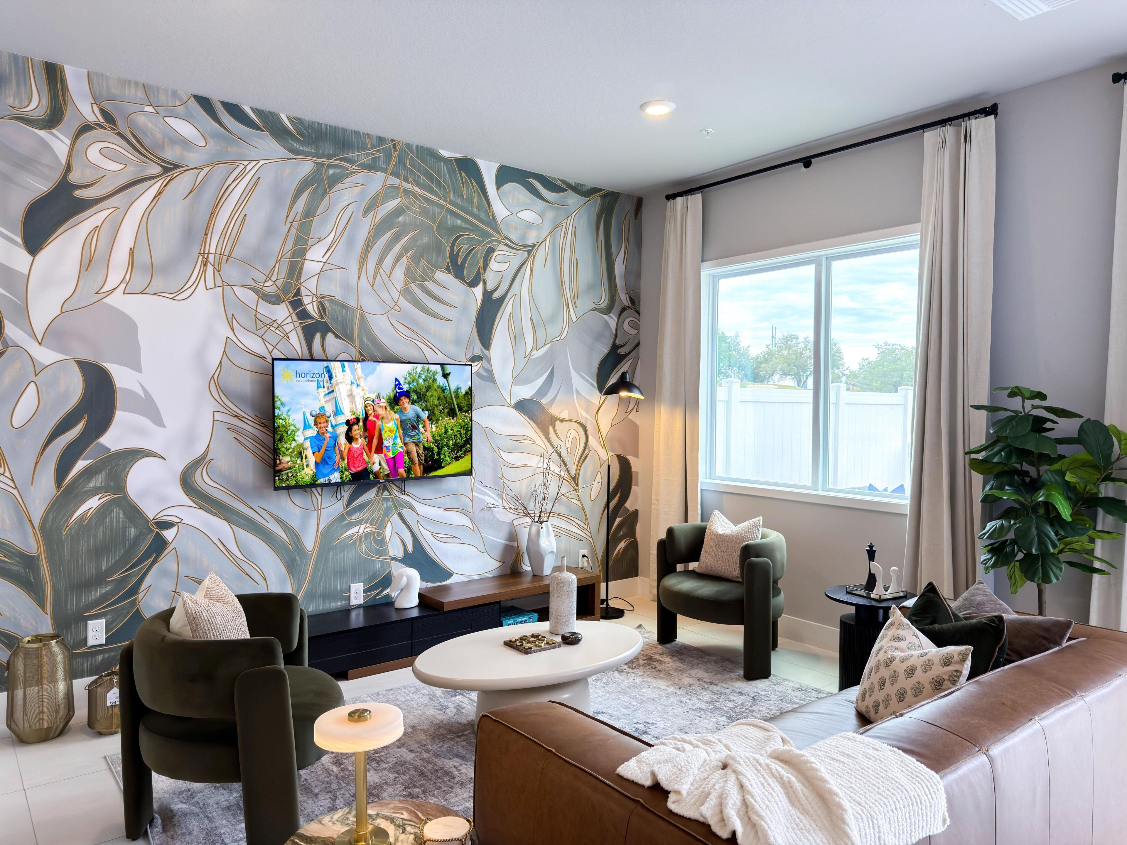 A modern living room with a large flat-screen TV mounted on the wall, a comfortable sofa, a coffee table, and a large window providing a view of the outdoors.