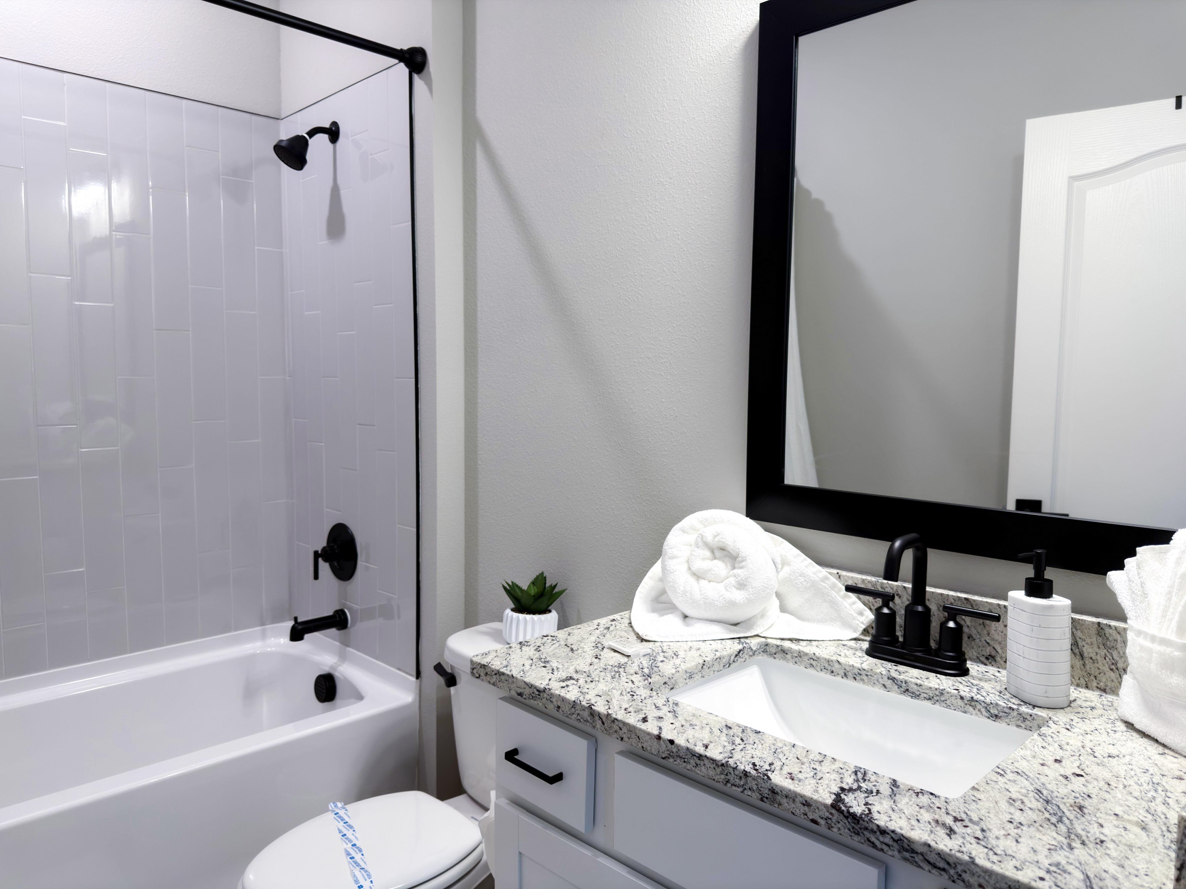 A modern bathroom with a white bathtub, a sink with a granite countertop, a small potted plant, and a mirror with a black frame.