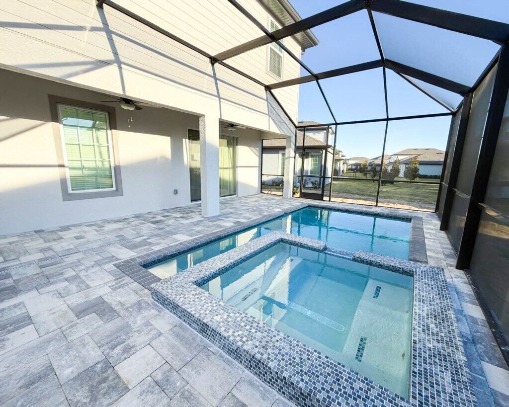 A swimming pool with a tiled deck, a covered area with a metal frame, and a view of a residential neighborhood.