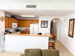 Una cocina moderna con armarios de madera, un refrigerador blanco y un microondas integrado. Hay una zona de comedor con una pequeña isla y sillas de mimbre.