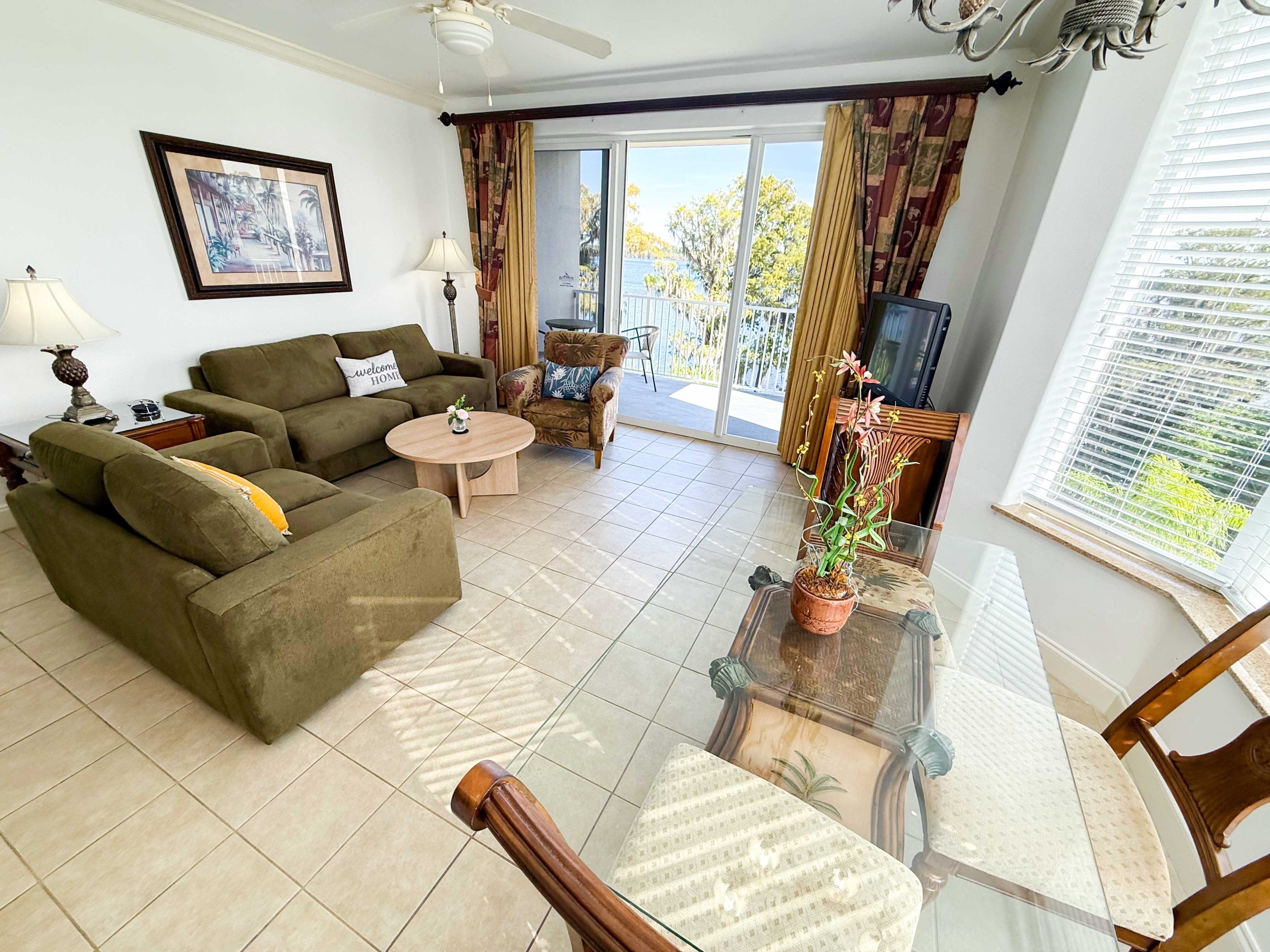 A living room with a sofa, a round coffee table, a glass-top table, and a chair. There is a balcony with a view of trees and a TV in the corner.