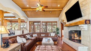 A spacious living room with a large sectional sofa, a fireplace, and a television mounted on the wall. - Rustic Retreat | 5 BR | Indoor & Outdoor Events (New Braunfels)