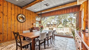 A wooden dining area with a large table and chairs, a hanging light fixture, and a view of the outdoors. - Rustic Retreat | 5 BR | Indoor & Outdoor Events (New Braunfels)