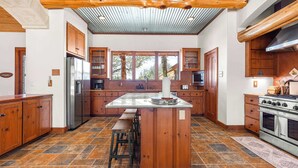 A spacious kitchen with wooden cabinets, a large island with stools, a stainless steel refrigerator, and a gas stove. - Rustic Retreat | 5 BR | Indoor & Outdoor Events (New Braunfels)