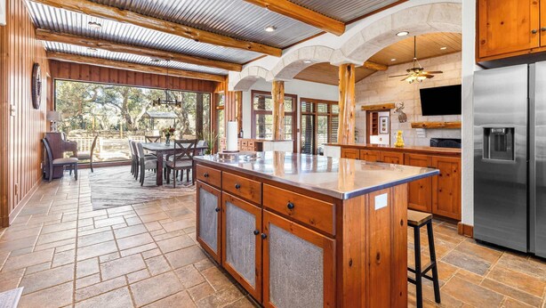 A spacious kitchen with wooden cabinetry, a large island, and a stainless steel refrigerator. - Rustic Retreat | 5 BR | Indoor & Outdoor Events (New Braunfels)