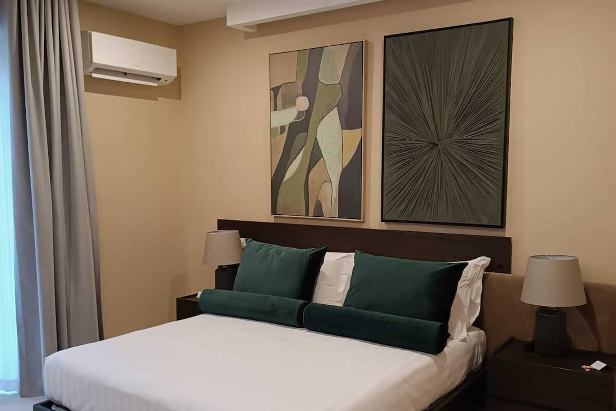 A modern hotel room with a large bed, two bedside tables with lamps, a wall-mounted air conditioner, and abstract artwork.