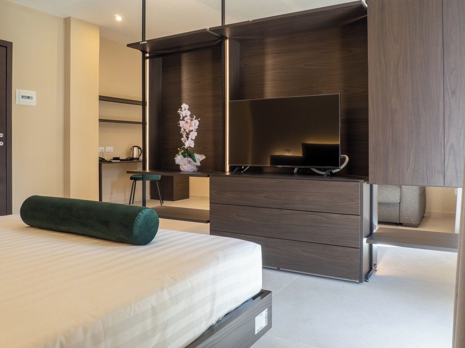 Un dormitorio moderno con una cama, una almohada cilíndrica verde, un aparador con cajones, un espejo y un pequeño escritorio con una silla.