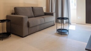 Ein modernes Wohnzimmer mit einem grauen Sofa, zwei schwarzen Beistelltischen und Blick auf ein Gebäude durch ein Fenster mit Vorhängen.