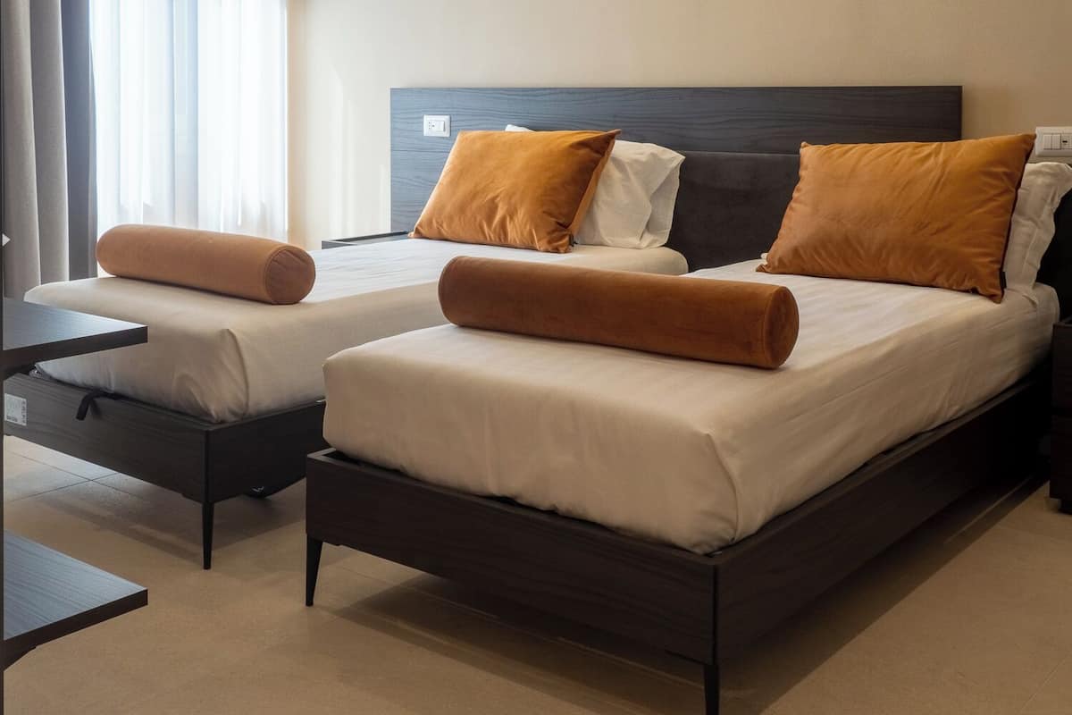 A hotel room with two beds, each with a dark headboard, white bedding, and orange pillows. There is a small table and a chair in the room.