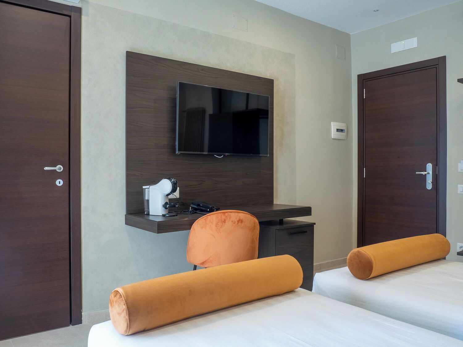 A hotel room with a bed, a desk with a chair, a television mounted on the wall, and two doors.