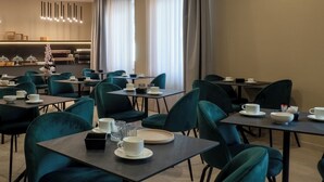 Ein Hotel-Essbereich mit mehreren Tischen und Stühlen, eine Küche im Hintergrund und große Fenster mit Vorhängen.