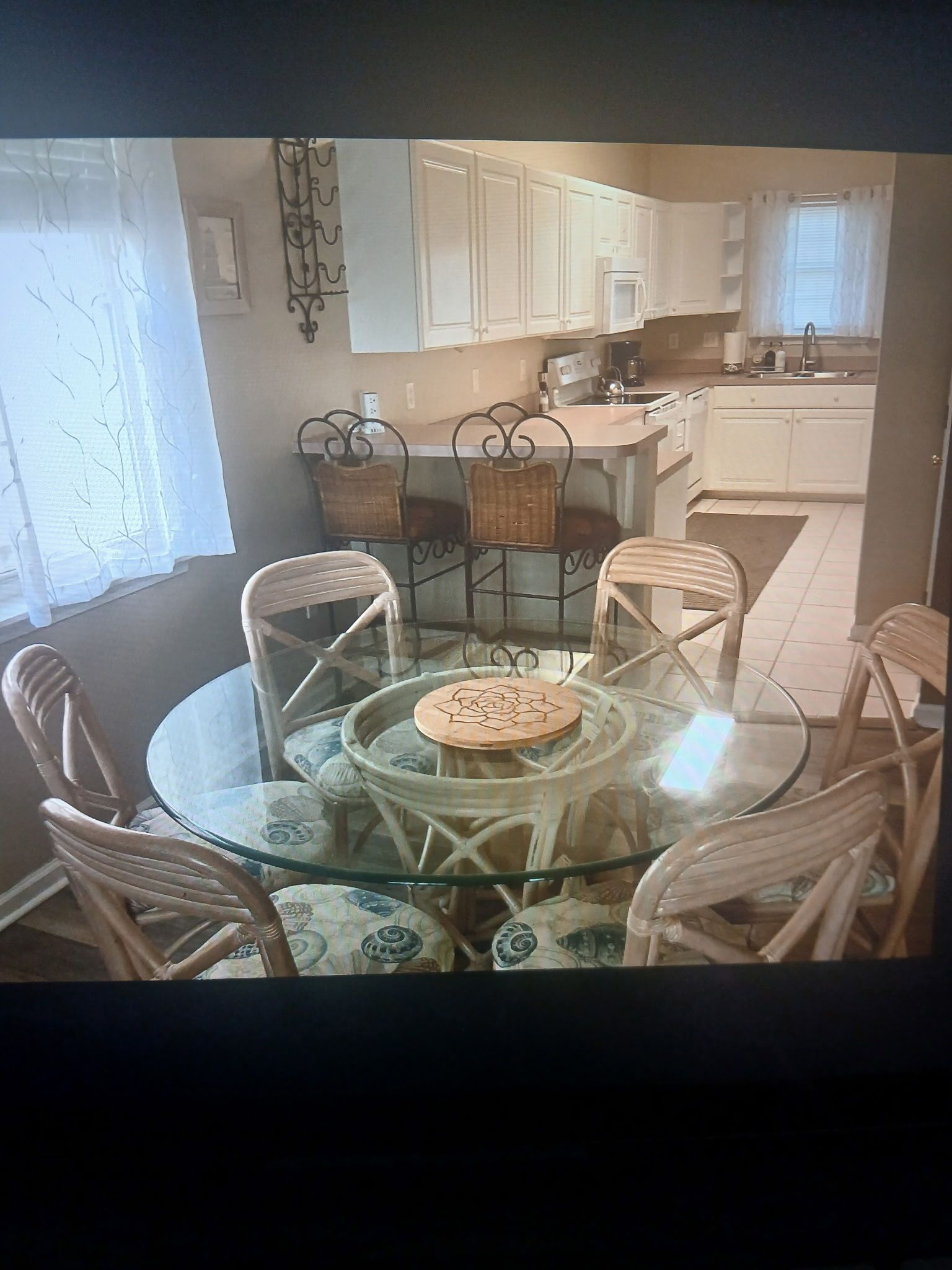 A dining area with a round glass table and wicker chairs, a kitchen in the background, and a window with a curtain.