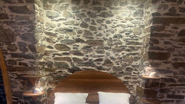 Ein Bett mit Holz-Kopfteil, weißer Bettwäsche und einer Wand aus Stein im Hintergrund.