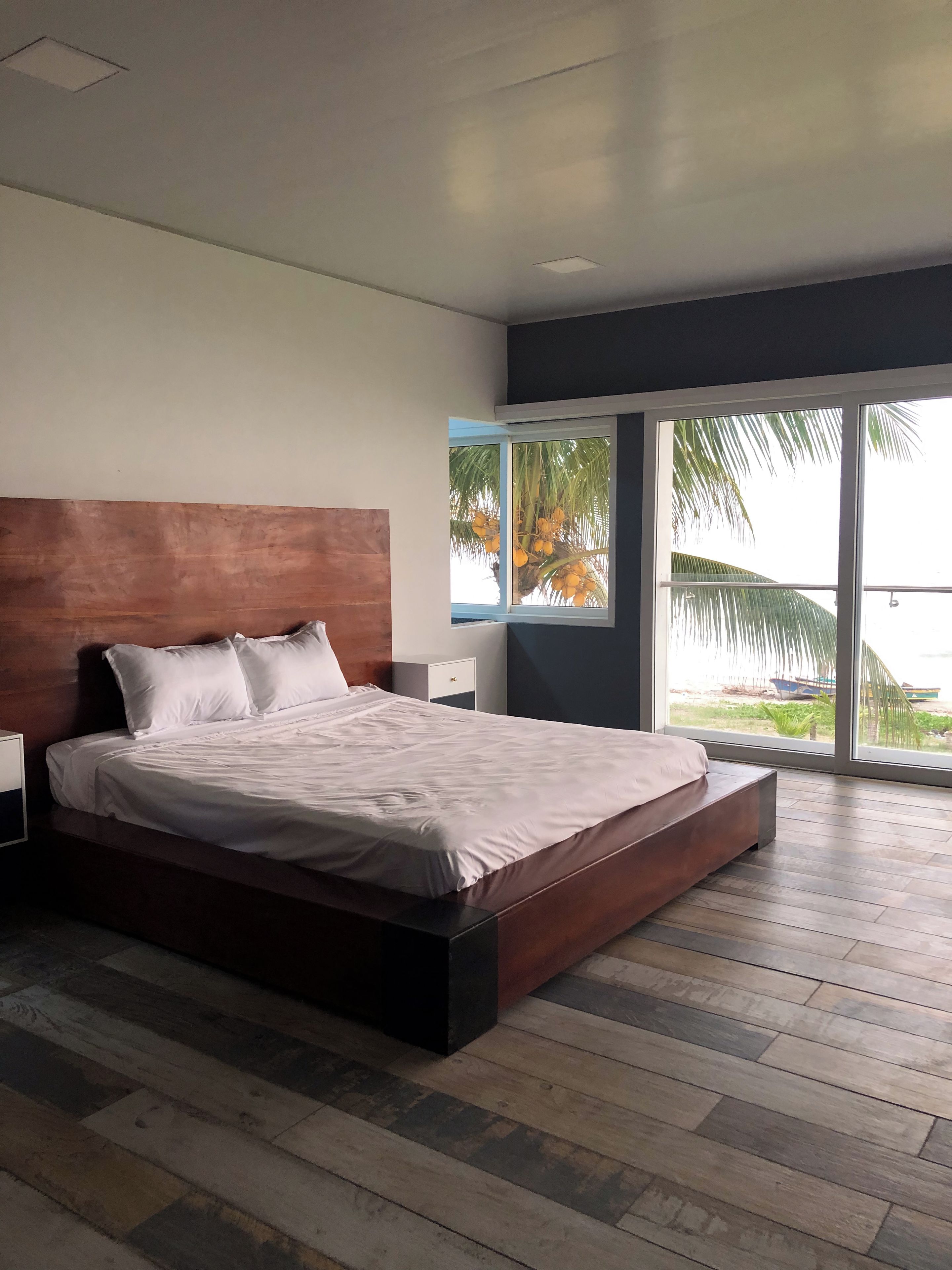 Een moderne slaapkamer met een groot bed, een houten hoofdbord en uitzicht op palmbomen door een groot raam.