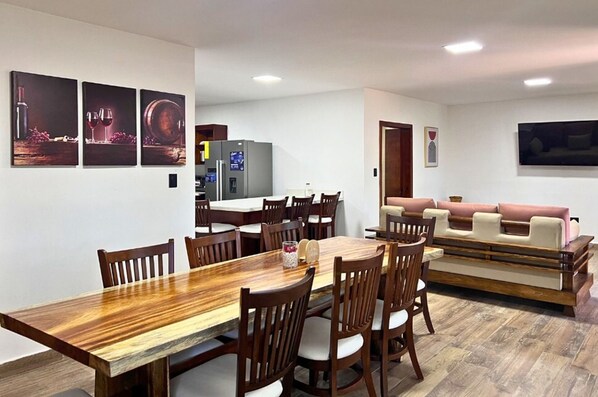 Un comedor con una mesa y sillas de madera, una cocina al fondo y una sala de estar con sofá y televisor.