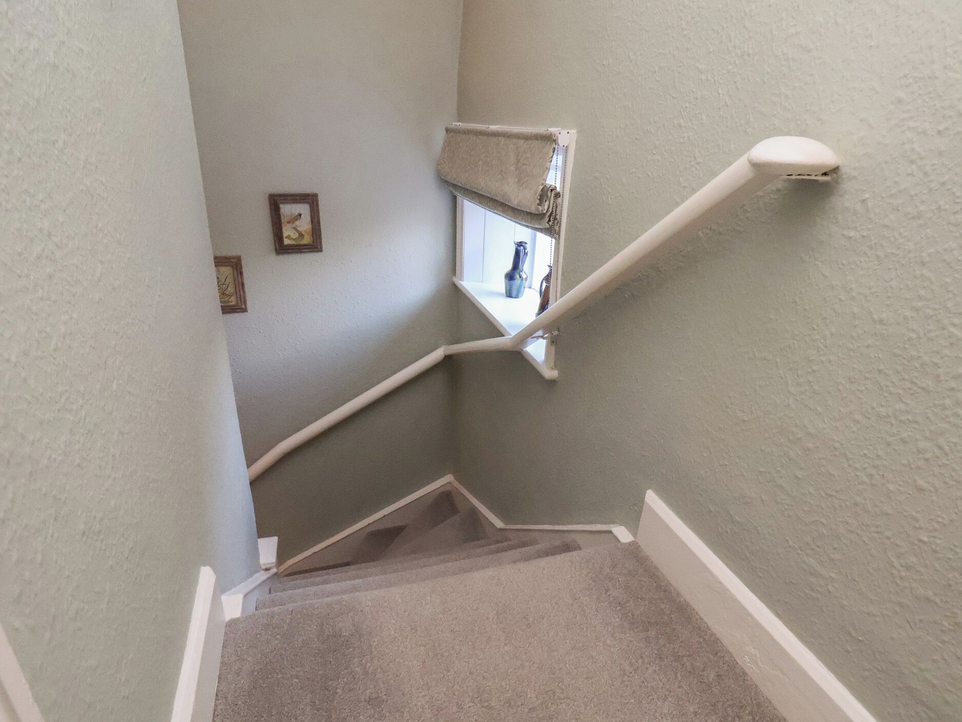 Un escalier recouvert de moquette, doté d’une main courante, d’une petite fenêtre et d’un cadre avec une image accroché au mur.