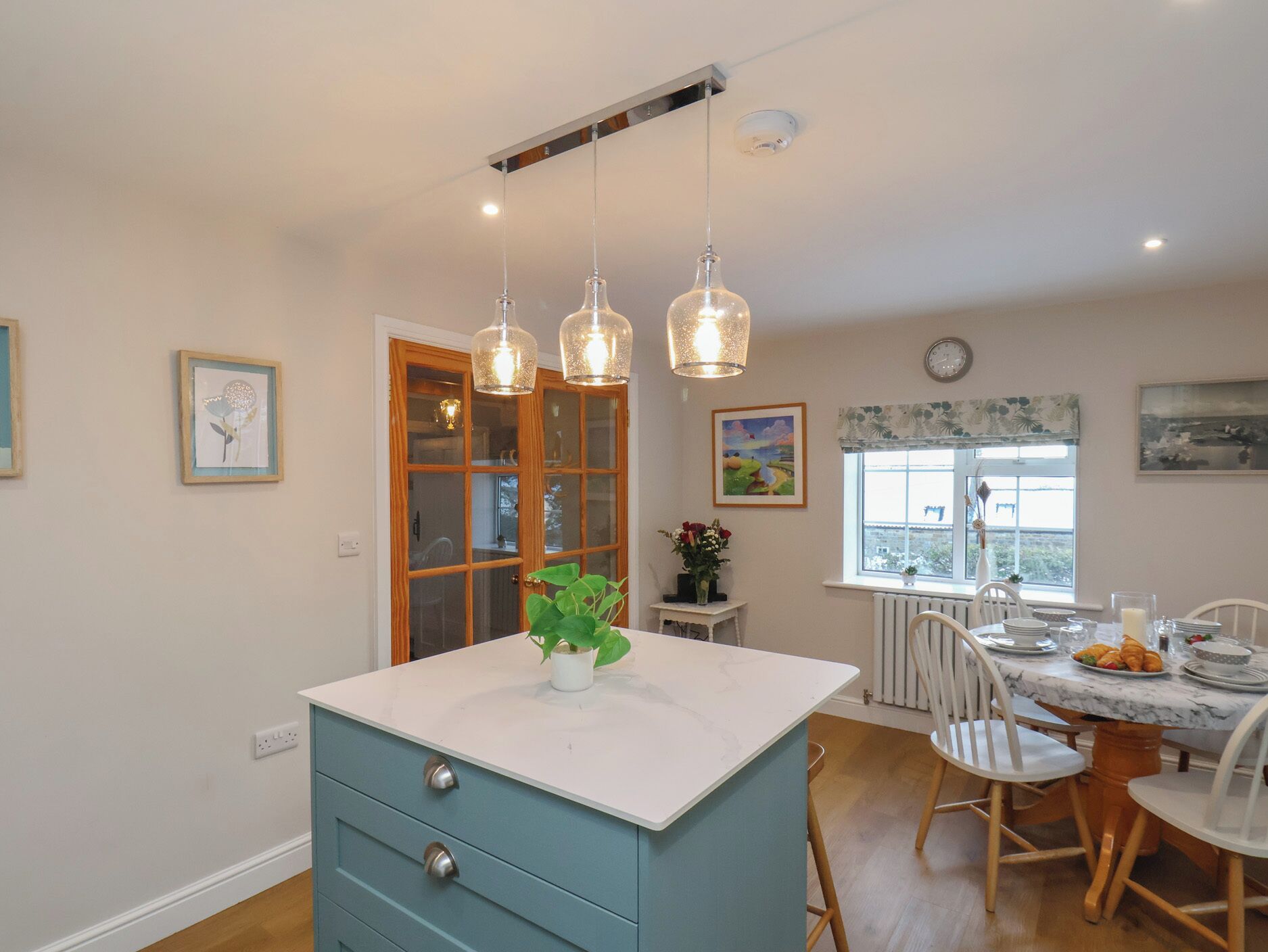 Une cuisine dotée d’un îlot central, de luminaires suspendus, d’un coin repas avec des chaises, d’une fenêtre offrant une vue et d’une horloge au mur.